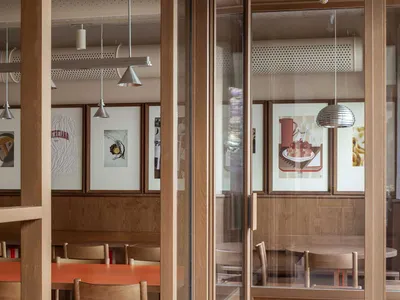 A modern restaurant interior with wooden tables and chairs, framed artwork, and industrial-style lighting.