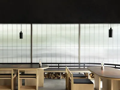 A modern restaurant interior featuring wooden furniture, glass elements, and a concrete floor.