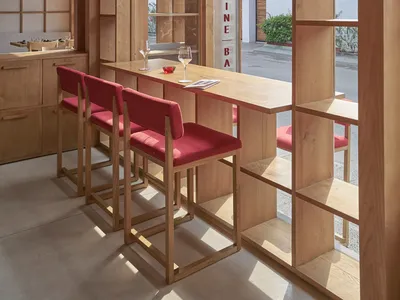 A modern restaurant interior featuring a wooden bar and seating area with red cushioned stools, large windows, and a minimali