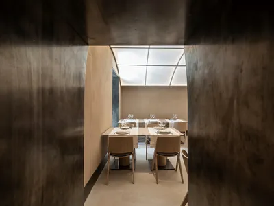 An empty, minimalist restaurant dining room with concrete walls, wooden furniture, and a skylight.