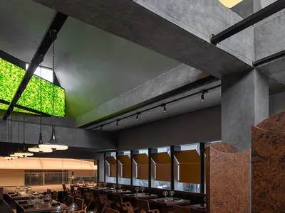 An industrial-style restaurant interior with concrete walls, wooden furniture, and a large green screen.