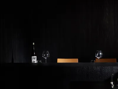 A minimalist restaurant interior with dark wood walls and tables, featuring a single bottle of sake and wine glasses, creatin