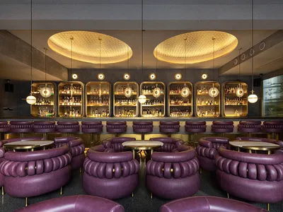 A modern restaurant bar area with purple leather chairs, glass shelves displaying bottles, and metallic pendant lights hangin