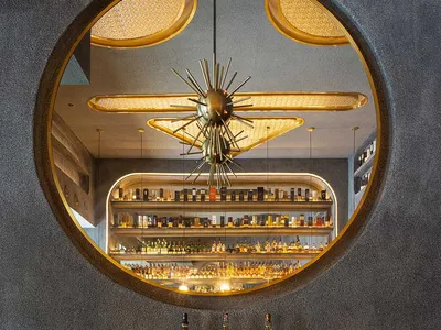 Modern industrial restaurant bar interior with concrete walls, brass accents, and circular window showcasing a well-stocked b