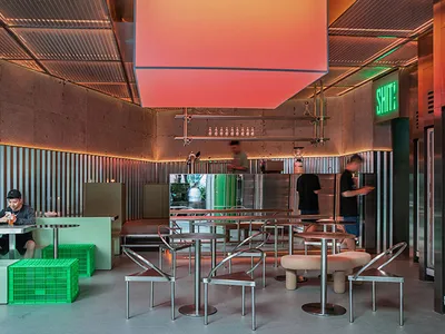 A modern restaurant interior featuring metallic tables, concrete walls, and green plastic baskets and chairs.