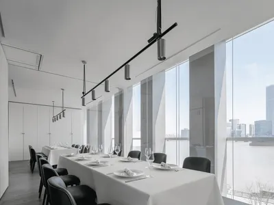 A minimalist restaurant interior with white walls and floors, featuring tables set with white tablecloths and black chairs.