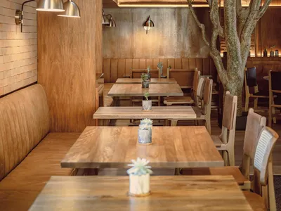 An industrial-style restaurant interior with wooden tables, wall-mounted lights, and a live tree in the center.