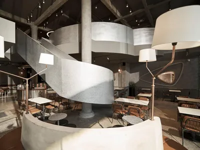 Concrete and steel industrial-style restaurant interior with spiral staircase and multiple seating areas.
