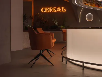A modern restaurant bar with concrete walls, illuminated by neon signs, featuring a stylish seating area with orange chairs a
