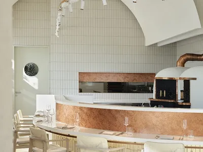 A minimalist restaurant bar with white tiled walls and concrete counter top, featuring bare wooden chairs.