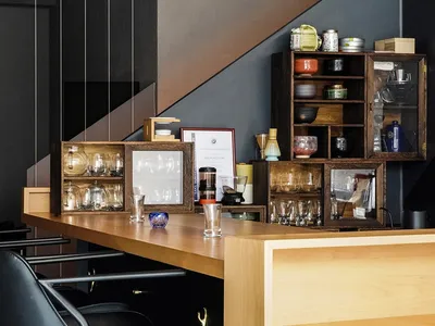 A modern restaurant bar interior featuring a wooden counter, metal stools, and various glassware and ceramic items on shelves