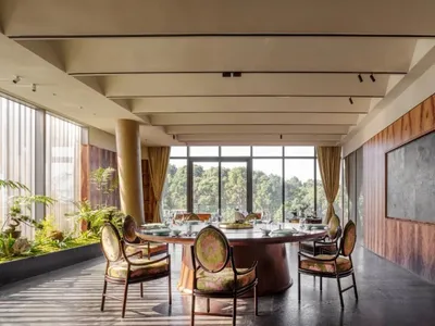 Modern restaurant dining room with round table, upholstered chairs, and large windows overlooking nature.