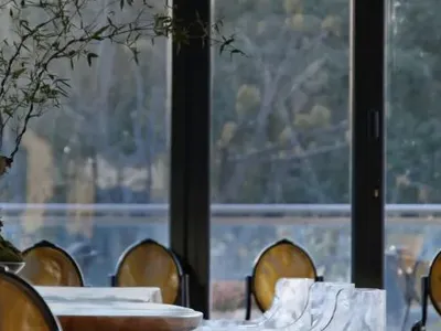 A modern restaurant interior featuring glass windows with a view of an outdoor green area and wooden tables.