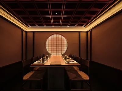 A modern restaurant interior with a long wooden table and chairs, illuminated by warm light from a circular window.