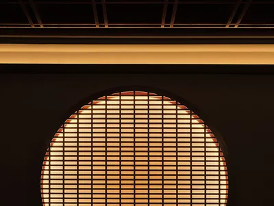 A long dark restaurant dining table with plates, chopsticks, and cups set for guests, with a large circular window in the bac
