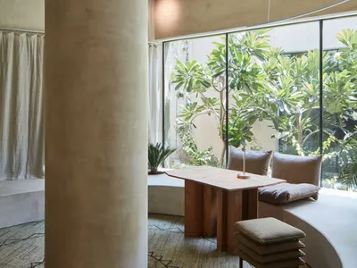 A modern home office with a natural wood table and modern seating area surrounded by large windows overlooking lush greenery.
