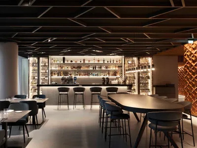 Modern restaurant interior with a bar and dining area featuring wood, concrete, and modern lighting fixtures.