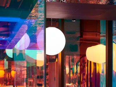 Modern restaurant interior with glass walls, illuminated tables and chairs, and colorful lighting effect.