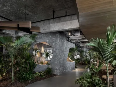 An industrial-style restaurant interior with concrete walls, wooden accents, and lush green plants.