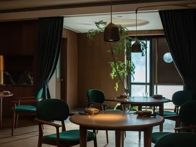 A modern restaurant interior featuring wooden tables, green fabric chairs, and glass accents.