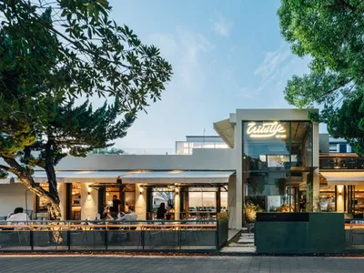 Outdoor seating area with modern restaurant exterior in white concrete and glass at dusk with trees and sky.
