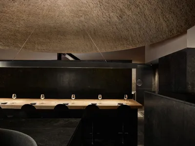 A minimalist restaurant interior with a long wooden table topped with wine glasses and plates, surrounded by black chairs and