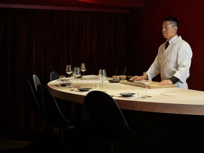 A chef stands at a modern restaurant counter with wood countertops and chairs.