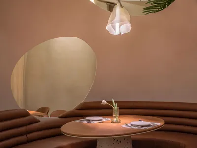 A modern restaurant interior with a curved leather bench seat and a table featuring a vase with a pink flower and cutlery.