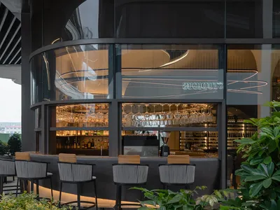 A modern restaurant bar featuring a glass partition, concrete furniture, and wine glasses hanging on the bar wall.