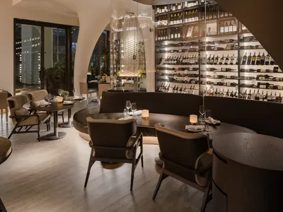 A modern restaurant interior with dark wood tables, glass wine shelves, and natural light through large windows.