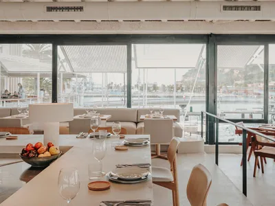 A modern restaurant interior with a long dining table, chairs, and a view of the marina through large windows.