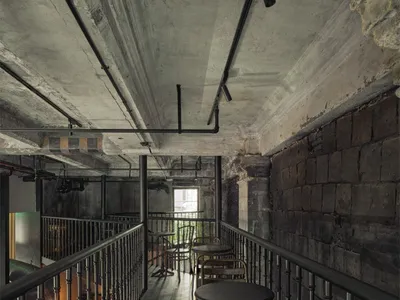 Industrial-style restaurant interior with concrete and brick walls, featuring metal railings and industrial lighting.