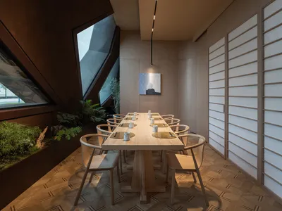 A modern restaurant interior featuring a long wooden dining table with chairs, surrounded by a combination of natural and man