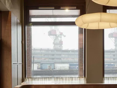 A modern restaurant interior with wooden tables, leather chairs, and large windows overlooking a harbor.