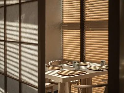 Restaurant interior with wooden chairs and blinds casting stripes on the wall.