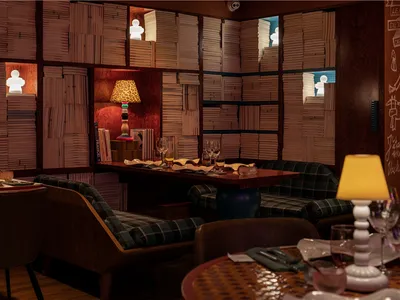 A modern restaurant bar library with wooden walls, paper books arranged on shelves, and a white lamp on a wooden table.