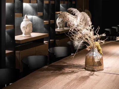 A modern restaurant interior with wooden accents and minimalist design, featuring Buddha statues and dried plants.