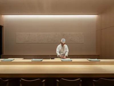 A chef in a traditional chef's hat prepares food behind a long wooden counter in a minimalist restaurant interior.