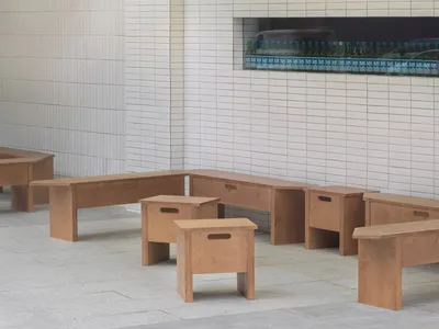 Modern restaurant interior with wooden benches and stools, white tiled walls, and a large window.