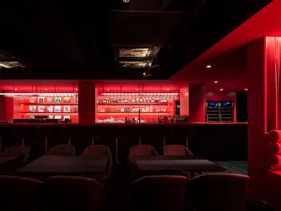A modern bar interior with red lighting and dark furniture.
