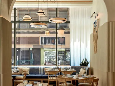 A modern restaurant interior with wooden tables, chairs, and floors, featuring a blue sofa with cushions, pendant lights, and