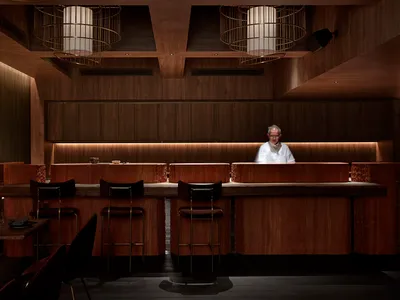 A modern restaurant bar with a wooden counter and stools, minimalist design, and warm lighting.