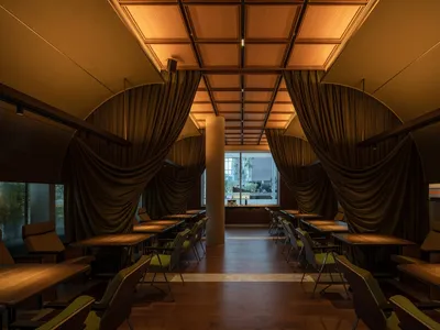 Modern restaurant interior featuring curtains and windows with a wooden floor and sustainable furniture.