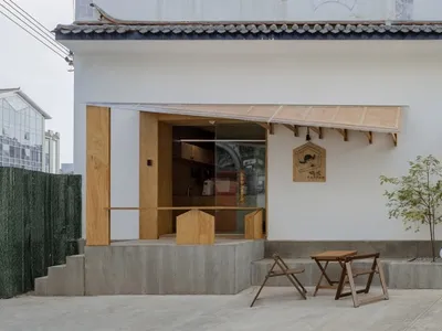 A minimalist façade with wooden accents and glass panels of a restaurant with a concrete base and a tree in front of it.