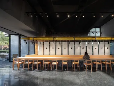 A modern restaurant interior featuring a long wooden counter with seating, minimalist black decor, and illuminated shelves.