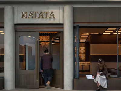 A casual-dining restaurant with a concrete exterior, featuring a person sitting in a window seat and another entering the est