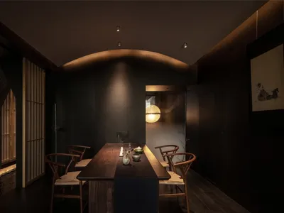 A minimalist dining room with dark wooden elements, featuring a long wooden table, chairs, and subtle lighting.