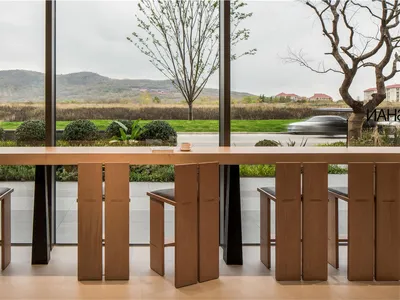 A café interior featuring a wooden counter and stools, with a view of a natural landscape.