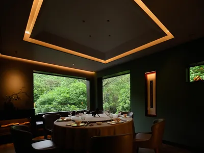 Modern restaurant interior with green leather seating and green glass windows showcasing lush greenery outside.