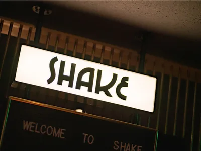 一塊霓虹燈招牌寫著「SHAKE」，懸掛在黑色邊框的留言板上方。